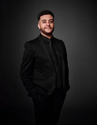 Mikael é um homem negro de pele clara, com cabelo preto e barba. Ele está usando uma camisa preta, um paletó preto e uma gravata preta com pequenas bolinhas brancas.