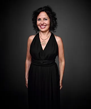 Patrícia é uma mulher branca, com cabelo preto, curto e cacheado. Ela está vestindo um vestido preto e um colar dourado.