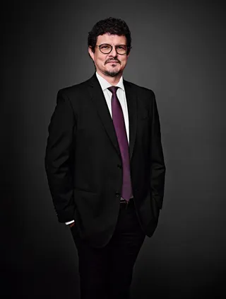 Sabah Teixeira é um homem branco, com cabelo preto curto e cacheado. Ele está usando uma camisa branca, uma gravata roxa e um blazer preto. Sabah usa óculos e tem um cavanhaque.